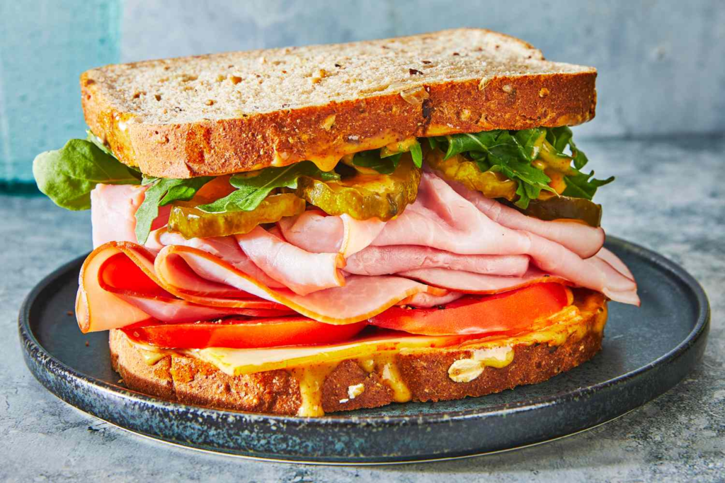 Loaded savory sandwich with sliced ham, cheese, tomato, pickles, and fresh lettuce layered between toasted whole-grain bread.