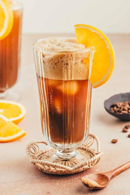 Refreshing iced coffee served in a tall glass, topped with creamy foam and garnished with a fresh orange slice.