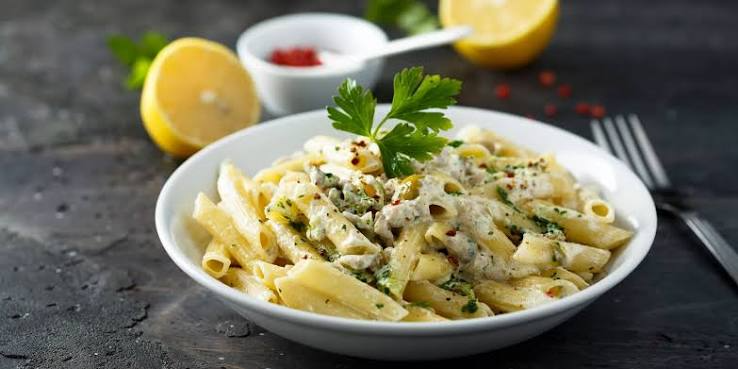 Corner Cafe creamy penne pasta with herbs, spices, and parsley garnish served in a white bowl with fresh lemon slices.